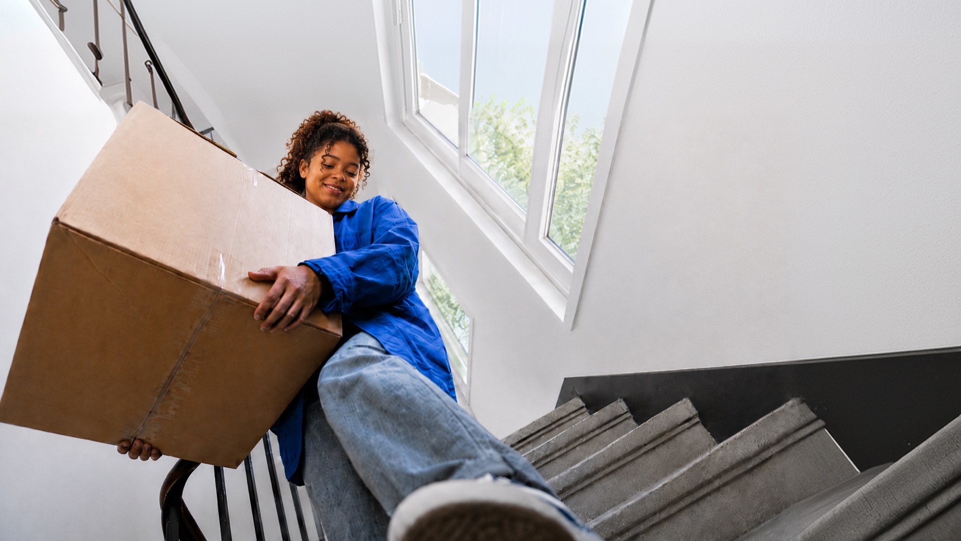 A woman in a blue shirt carrying a large cardboard box down a stairwell