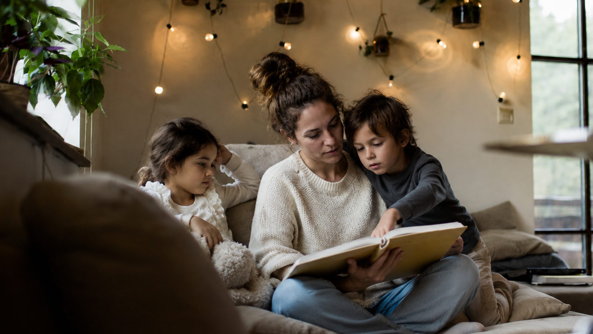 A mother reading a picture book with two children by string lights